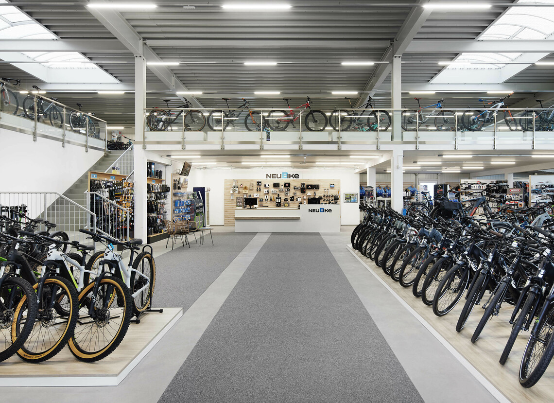 Innenansicht der NEUBIKE Stores: großes Fahrradsortiment auf zwei offenen Etagen; eine graue Teststrecke führt geradeaus zum Verkaufstresen; die Einrichtung ist hell, modern und freundlich mit hellen Holzelementen.