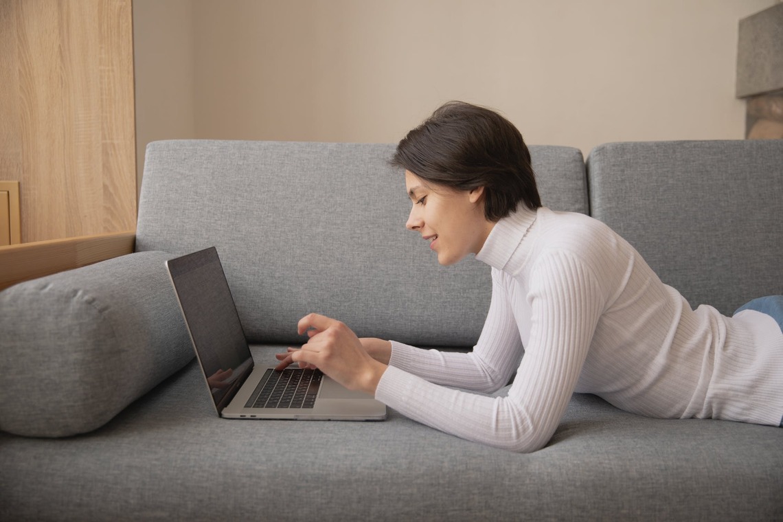 Frau liegt auf dem Sofa und tippt etwas in ihren Laptop ein. 