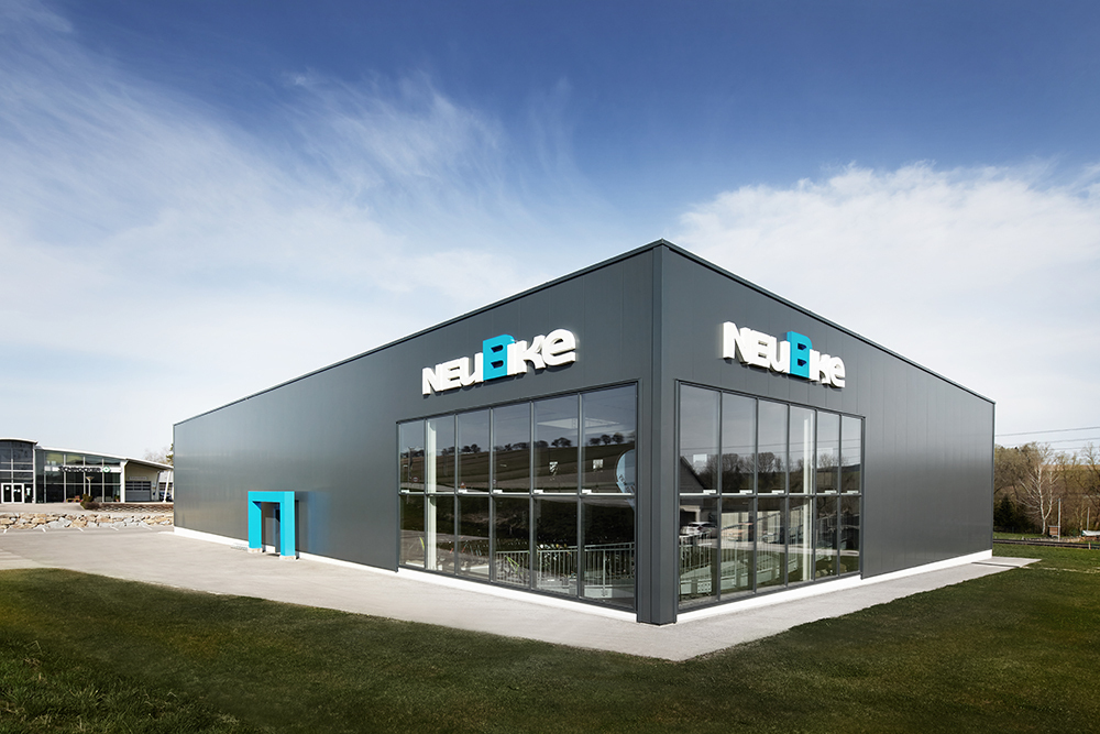 Außenansicht des NEUBIKE Stores in Grieskirchen; dunkelgraue Fassade mit zwei großen Schaufensterfronten, über denen das NEUBIKE-Logo platziert ist. Ein blauer Türrahmen lädt zum Eintreten ein.